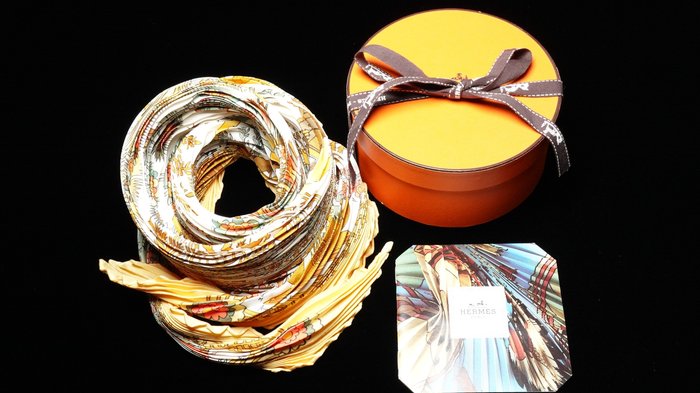 A folded patterned scarf displayed beside an orange oval gift box with a brown ribbon and a small matching card, all photographed on a black background.