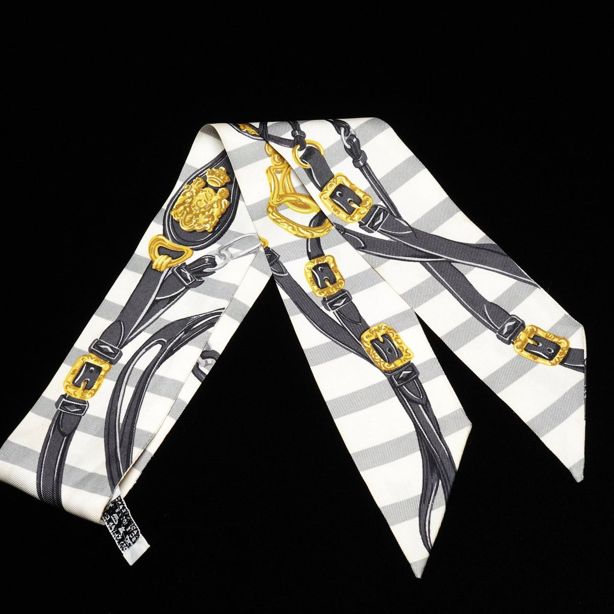 A silk scarf featuring a harness and bit pattern with black straps, gold buckles, and a white and gray striped background, displayed against a solid black backdrop.
