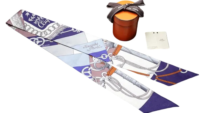 An Hermes silk scarf with a purple, white, and orange horsebit design, displayed alongside an orange Hermes gift box and a white paper tag on a black background.