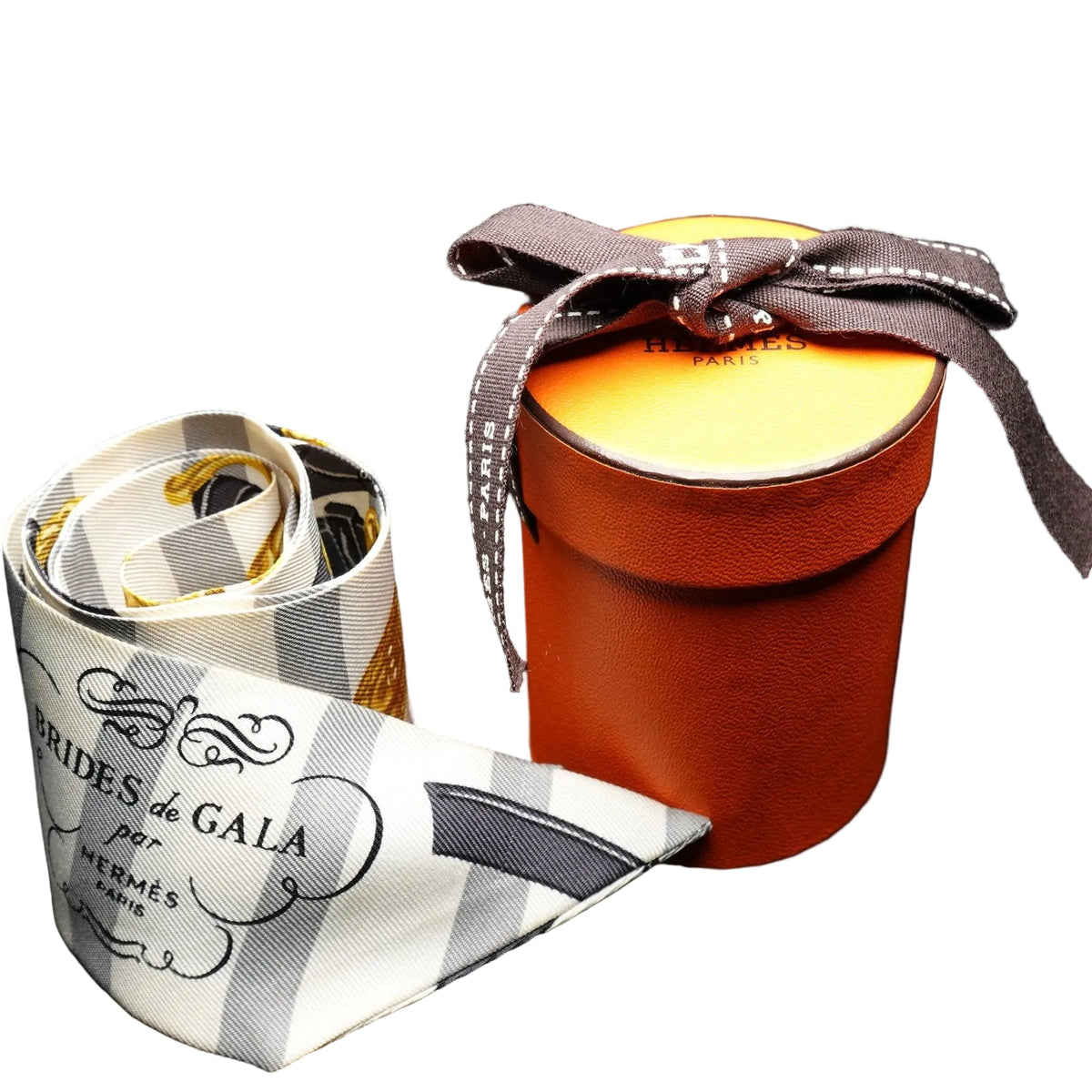 A Hermes-branded scarf with a white, gray, and black check pattern and gold trim, displayed next to its signature orange cylindrical box with a brown ribbon bow on a black background.