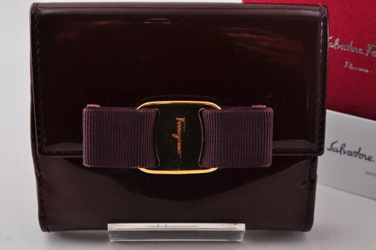 Salvatore Ferragamo glossy dark burgundy patent leather wallet with a signature bow tie closure featuring a gold-tone buckle and gold logo.